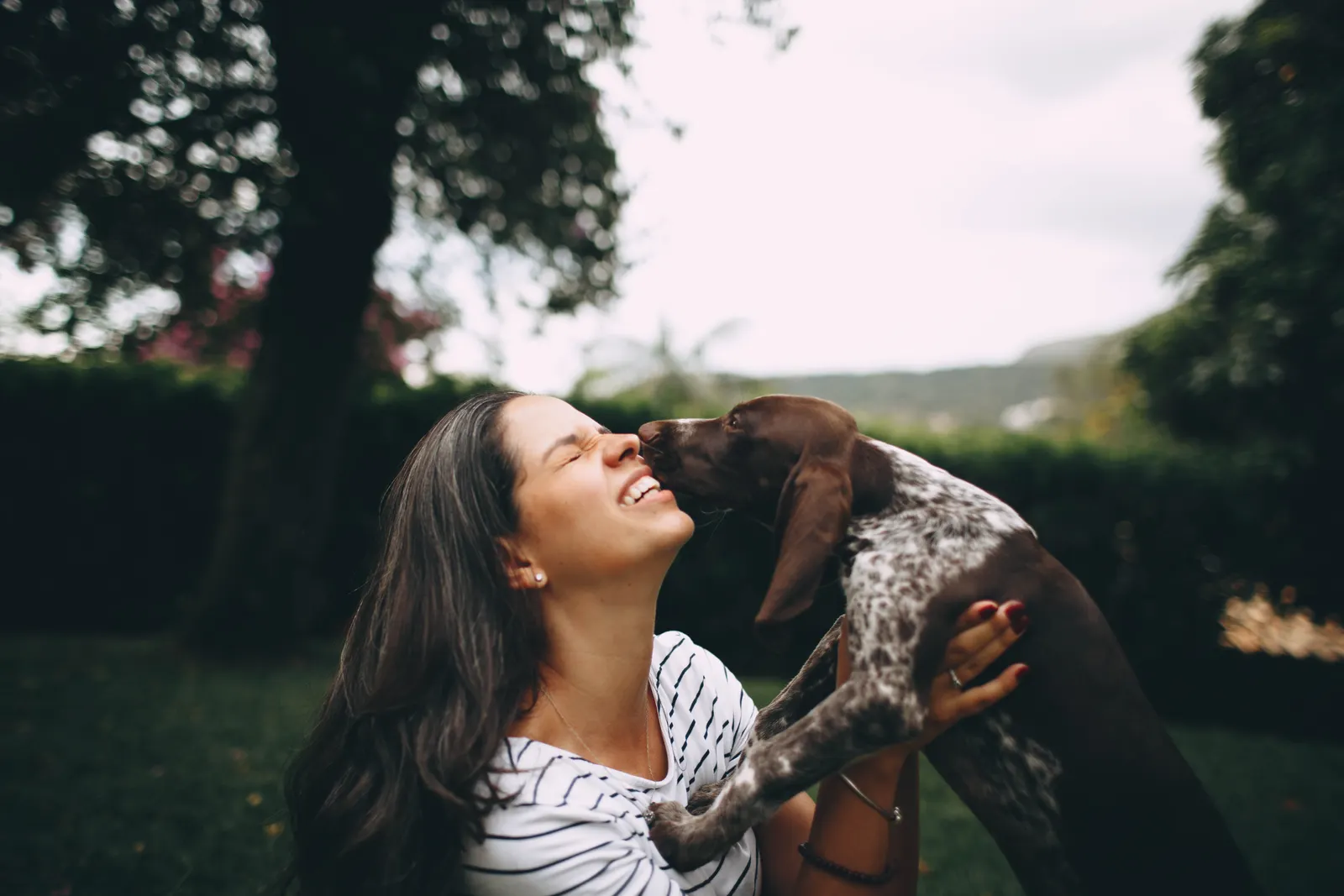 Un chien brun et blanc lèche le visage d’une femme qui le tient dans ses bras, dans un jardin.